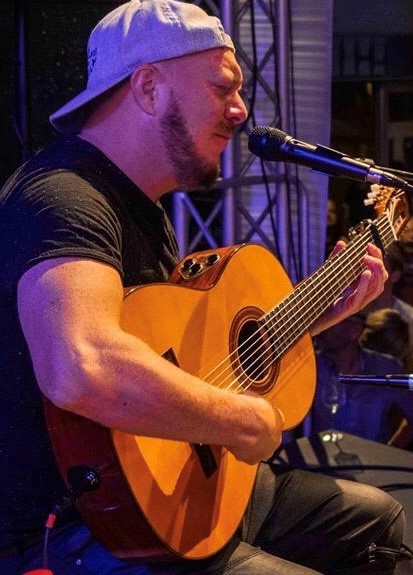 romero Man with beard wearing a baseball cap backwards is playing an acoustic guitar and singing into a microphone.