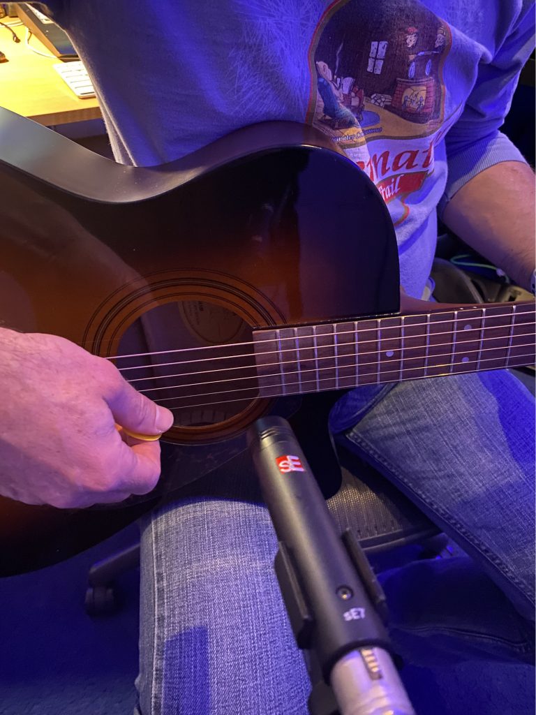 Recording-Acoustic-Body-image-1 Closeup of someone's hands as they play acoustic guitar with a small directional microphone positioned directly at strings.